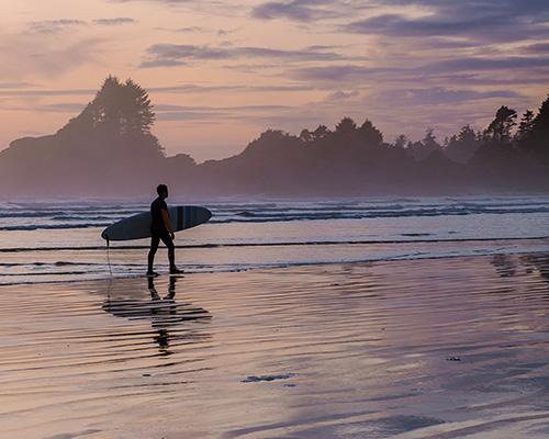 A figure carrying a surfboard on the beach in sunset.