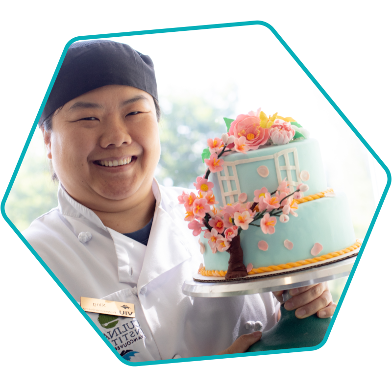 Smiling woman in baker's whites holding a fancy cake. 